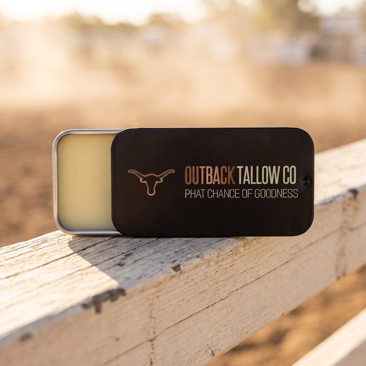 The OG tallow balm slide tin open on a weathered rodeo arena rail (AI-generated image)
