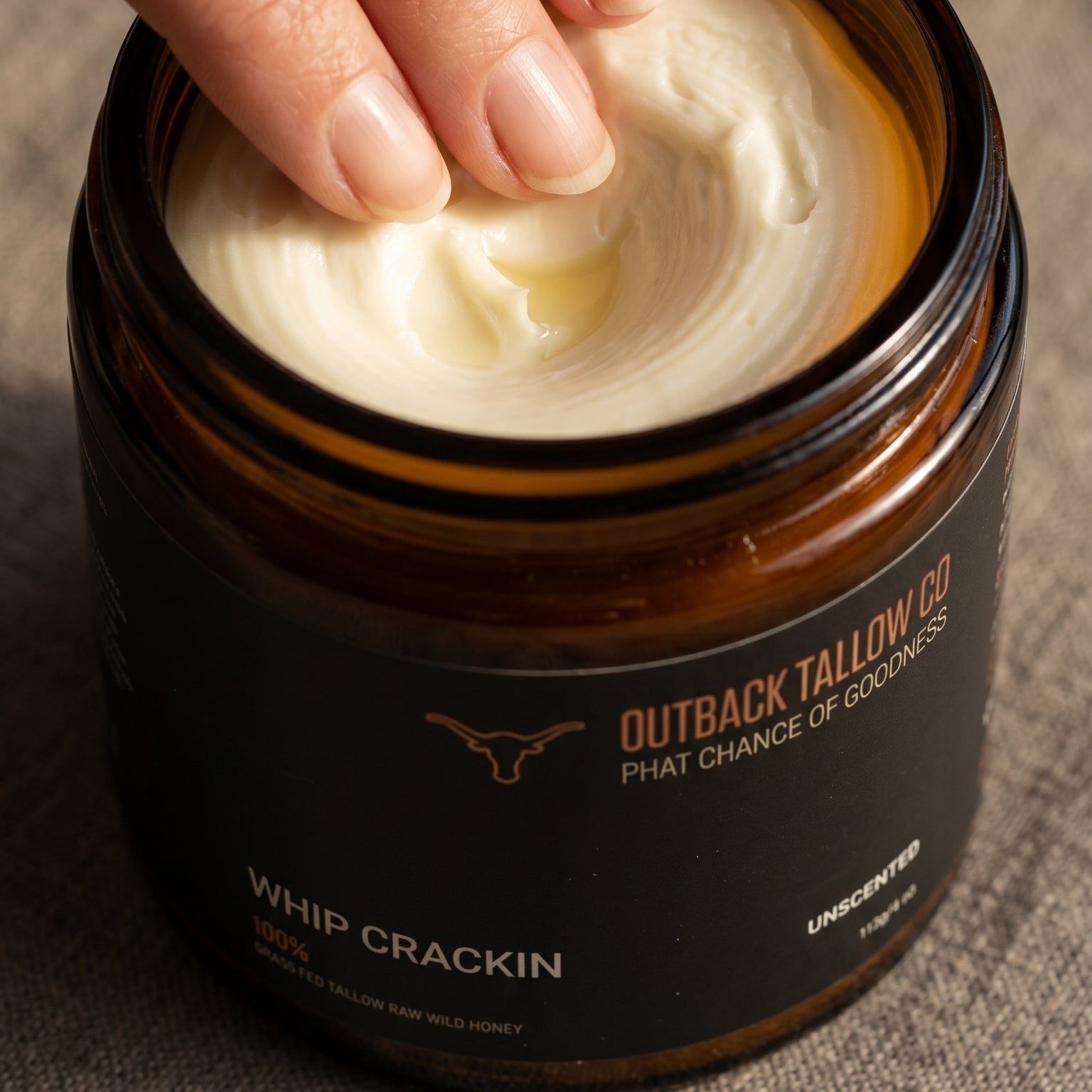 Hand reaching into a jar of Outback tallowco Co whip crackin phat balm on a textured surface.