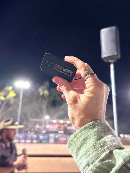 Hand holding The OG tallow balm slide tin up at a rodeo at night