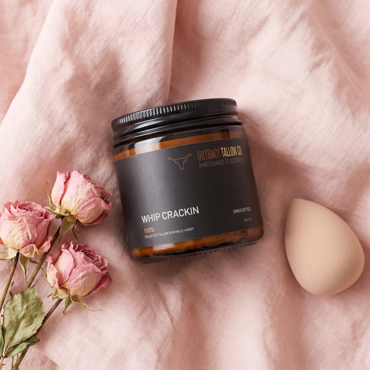 Whip Crackin' whipped body moisturiser jar on blush pink linen with dried roses (AI-generated image)
