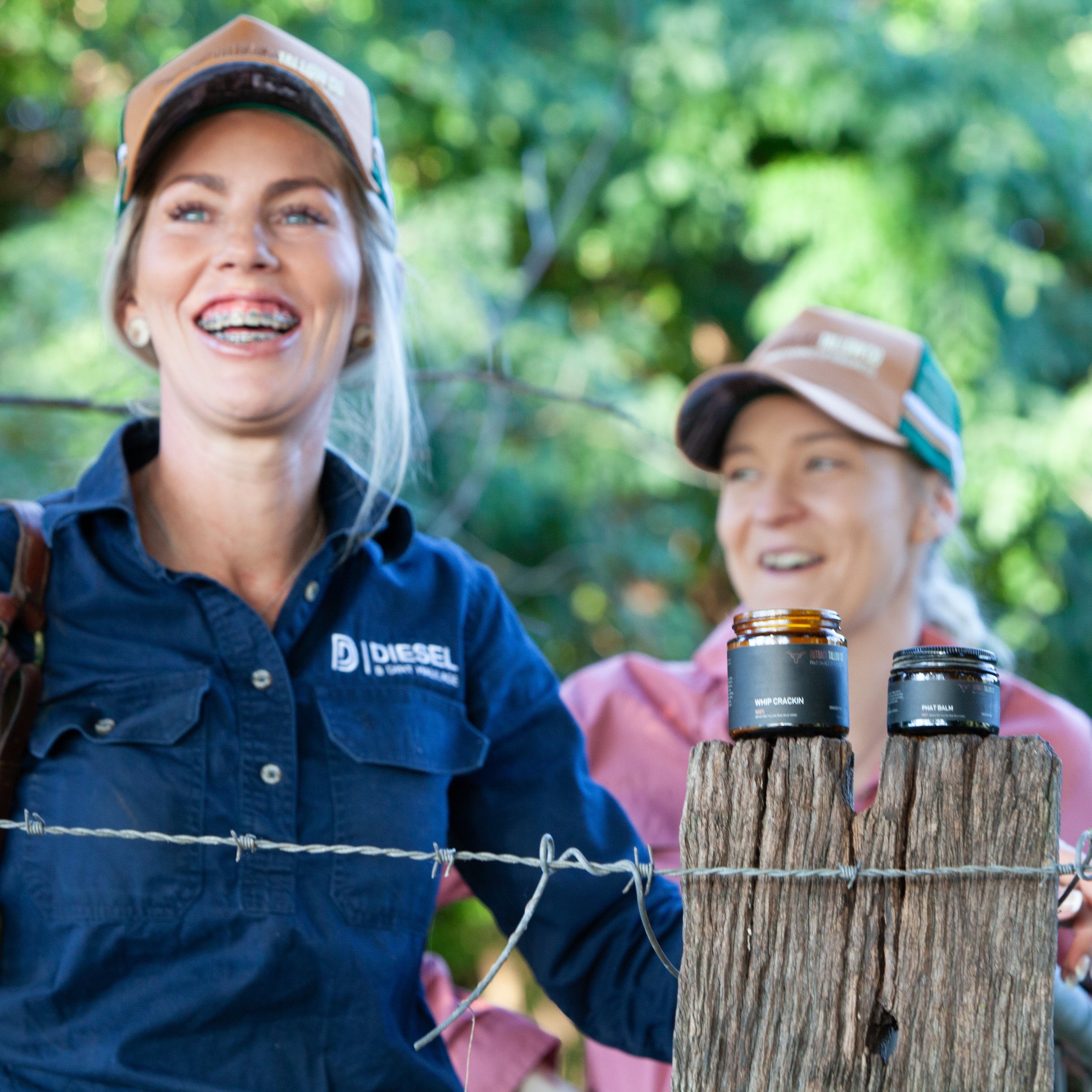 Two women standing behind a barbed wire fence with beef tallow on a log, surrounded by greenery.